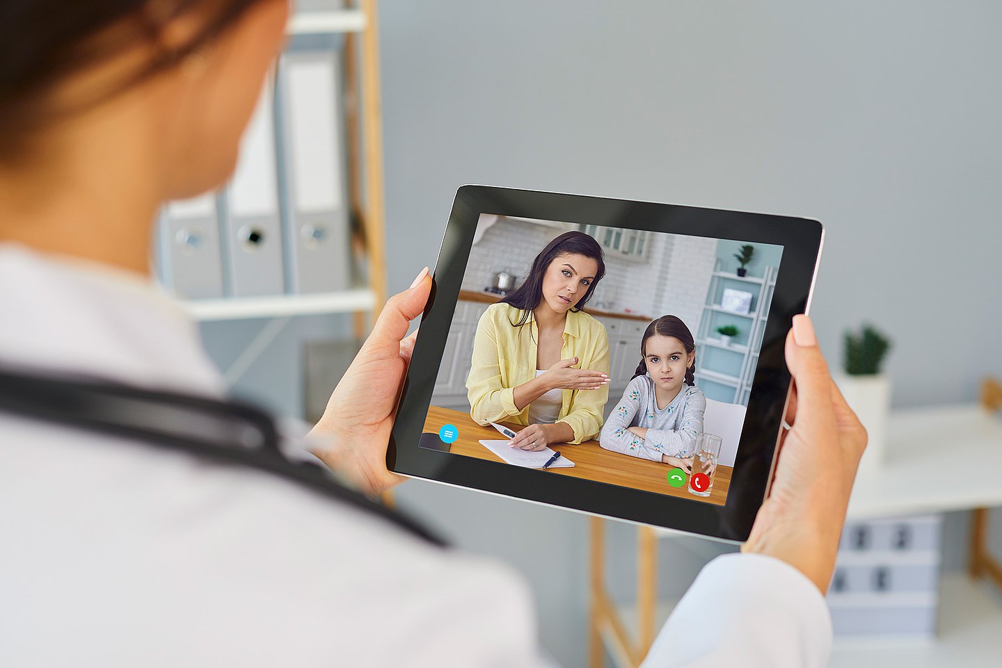 Eine Ärztin führt eine Videosprechstunde mit einer Patientin und deren Tochter durch. Die beiden sind auf dem Tablet der Ärztin sichtbar, die in ihrem Sprechzimmer sitzt.