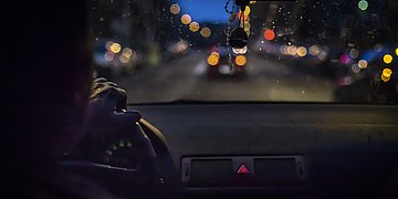Foto: Aus der Perspektive vom Hintersitz eines Autor sitzt eine Person am Steuer. Eine Hand ist am Lenker. Im Fenster baumeln Anhänger. Draußen ist es Nacht und die Straße in der Stadt ist beleuchtet.