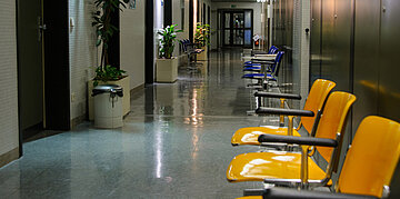 In einem menschenleeren Krankenhausflur stehen links an der Wand Pflanzenk&uuml;bel zwischen den T&uuml;ren und rechts sind mehrere gelbe und blaue Plastikst&uuml;hle aufgestellt.