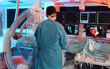 Herzkatheter-Labor Foto zeigt einen Arzt im grünen OP-Kittel mit dem Rücken zu Betrachter, auf dem OP-Tisch eine Patientin, einghüllt in Schutzfolie, der ein Herzkatheter gelegt wird. Im Hintergrund Computermonitore. über die der Arzt den Eingriff überwacht.a