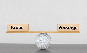 Weltkrebstag 2025 Schleswig-Holstein Mit einem kleinen Ball und einem Holzbrett mit den Gewichten 'Krebs' und 'Vorsorge' wird eine Waage dargestellt, die im Gleichgewicht ist.