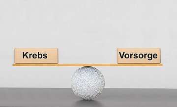 Mit einem kleinen Ball und einem Holzbrett mit den Gewichten 'Krebs' und 'Vorsorge' wird eine Waage dargestellt, die im Gleichgewicht ist.