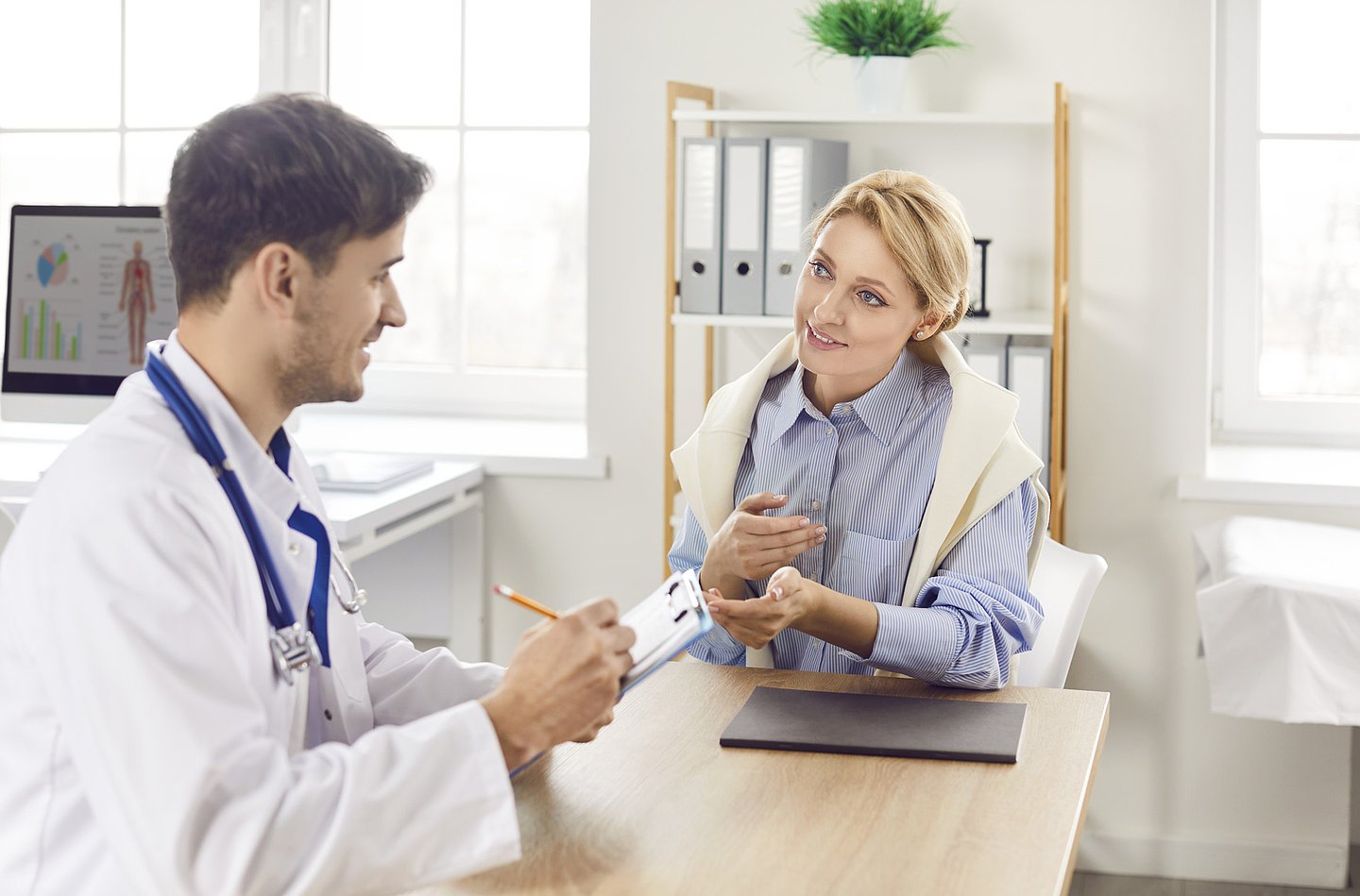 Ein Arzt und eine Patientin sitzen im Behandlungszimmer, sie sind in einem Gespräch vertieft. Der Arzt notiert etwas auf einem Formular, das er (Klemmbrett) in der Hand hält.