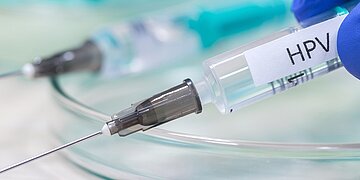 eine Spritze mit der Aufschrift "HPV", im Hintergrund Laborutensilen