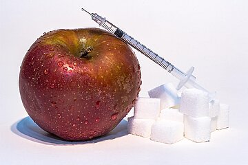 Symbolfoto zum Thema Diabetes, das einen Apfel, eine Insulinspritze und einen Stapel Würfelzucker zeigt.