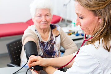 Eine &Auml;rzte sitzt am Tisch eines Behandlungszimmers und misst bei einer &auml;lteren Patientin den Blutdruck.