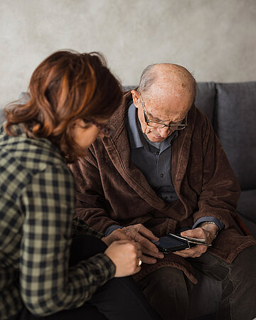 Foto: Eine Frau sitzt neben einem &auml;lteren Mann auf einem Sofa und unterst&uuml;tzt ihn beim Blick auf ein Smartphone.