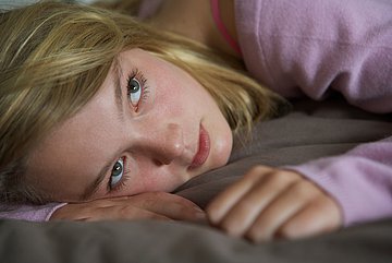 Long-Covid in Westfalen-Lippe Foto zeigt das Gesicht einer jungen Frau, die auf dem Bauch liegend, mit offenen Augen in Blickrichtung schaut.