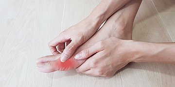 Gicht Eine Person fasst sich an das Gelenk des großen Zehs, welches gerötet ist.