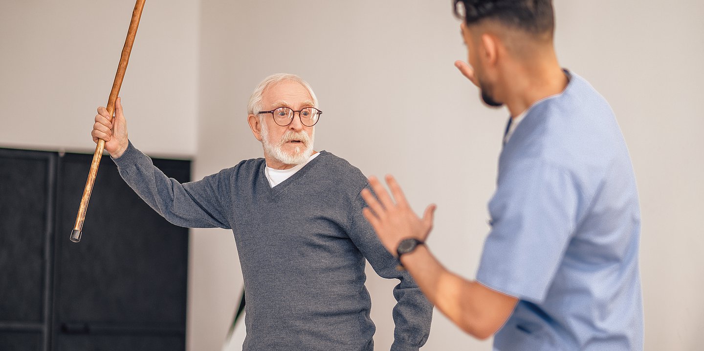 Ein älterer Mann erhebt seinen Gehstock gegen einen jüngeren Mann in blauer Pflegekleidung.