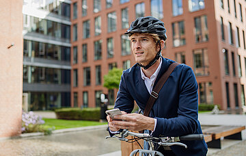 Ein junger Mann steht mit seinem Fahrrad vor weinem B&uuml;rogeb&auml;ude. Er st&uuml;tzt sich sich auf den Lenker und h&auml;lt ein Smartphone in seinen H&auml;nden.Er tr&auml;gt einen Fahrradhelm, ein dunkelblaues Sakko, darunter ein blasslaues Oberhemd. Um den Rumpf liegt der braune Lederriemen seiner Aktentasche. Im Hintergrund sind vor dem B&uuml;rogeb&auml;ude B&auml;ume, eine Hecke und ein Rasen zu sehen.