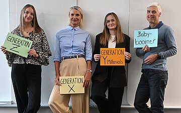 Vier Personen aus vier Genrationen stehen nebeneinander und halten Schilder f&uuml;r die Generationen X, Y und Z sowie f&uuml;r "Babyboomer" in H&auml;nden.