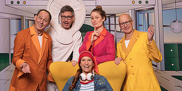 Auf einem gelben Sessel sitzt eine kost&uuml;mierte Frau. Hinter dem Sessel stehen vier weitere Personen in bunten Anz&uuml;gen, der eine von ihnen tr&auml;gt ein Kochl&ouml;ffelkost&uuml;m.