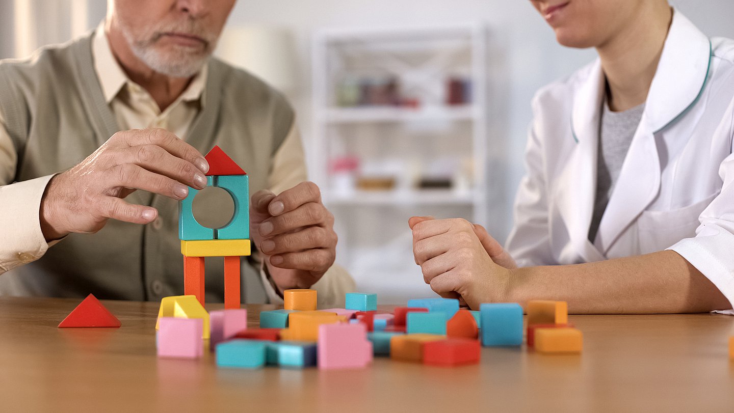 Foto eines pflegebedürftigen Mannes, der an einem Tisch sitzt und mit farbigen Bauklötzen, die unterschiedlich geformt sind, einen Turm baut. An seiner Seite sitzt eine Therapeutin, die ihn dabei unterstützt.