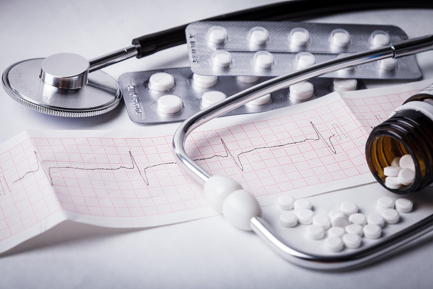 Foto zeigt ein Stethoskop, einen Ausdruck vom EKG, Tablettenblister sowie eine offenen, umgekippten Behälter, aus dem Tabletten herausgefallen sind.