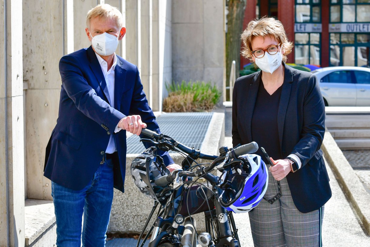 Foto: Dr. Jürgen Peter und Daniela Behrens stehen vor einem Gebäude nebeneinander. Sie halten ihre Fahrräder, die zwischen ihnen stehen und tragen Atemschutzmasken. 