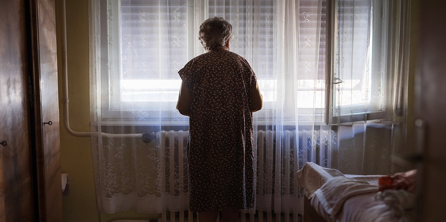 Foto: Eine alte Dame steht im abgedunkelten Zimmer und schaut aus dem Fenster, vor dem aber ein Rollo runtergefahren ist.