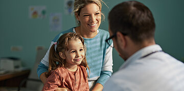 Mutter sitzt mit ihrer Tochter in einer Arztpraxis und spricht mit einem Arzt.