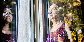 An einem Fenster steht eine &auml;ltere Dame. Die Ansicht wird in der Scheibe gespiegelt.