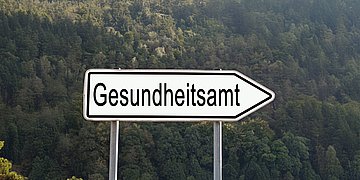Foto: Ein weißes Straßenschild zeigt nach rechts, darauf der Schriftzug "Gesundheitsamt".