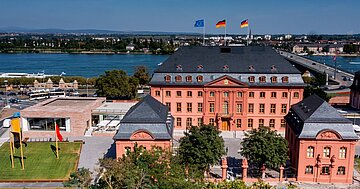 Foto zeigt den Blick auf das historische rotbraune Geb&auml;ude im Hintergrund flie&szlig;t der Rhein
