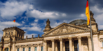 Das Reichstagsgeb&auml;ude in Berlin mit wehenden deutschen Flaggen unter einem teils bew&ouml;lkten Himmel.