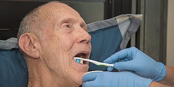 Mundhygiene in der Pflege Foto zeigt einen Senior mit Glatze und zwei Hände mit blauen Einmalhandschuhen. Dem Mann werden die Zähne geputzt.