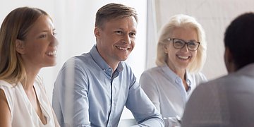 Selbsthilfefreundlichkeit Foto von zwei Frauen, die mit einem Mann in der Mitte einem weiteren Mann am Tisch gegenübersitzen, der nur von hinten zu sehen ist. Sie sehen gut gelaunt aus.