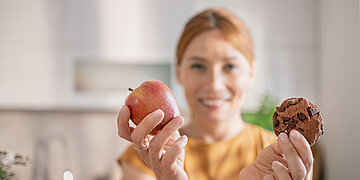 Eine l&auml;chelnde Frau h&auml;lt einen Apfel und einen Schokoladenkeks in die Kamera; die beiden Lebensmittel sind scharf im Vordergrund, die Frau und die K&uuml;che im Hintergrund unscharf.