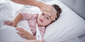 Ein Kleinkind liegt im Bett, die Mutter h&auml;lt eine Hand auf den Kopf des Kindes und ein Fieberthermometer in der anderen Hand.