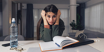 Foto: Ein Frau sitzt am Tisch &uuml;ber einem aufgeschlagenem Buch und guckt frustriert.