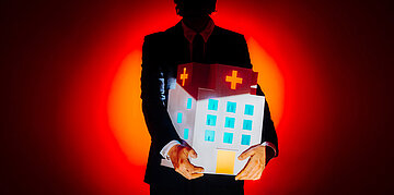 Eine schattenartige Person steht vor rotleuchtendem Hintergrund. Im Arm tr&auml;gt sie ein Krankenhaus.