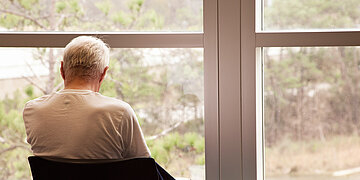 Ein Mann sitzt vor dem Fenster und schaut raus. Das Foto ist von hinten aufgenommen.