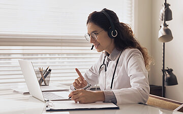 Foto: &Auml;rztin sitzt vor einem Laptop und tr&auml;gt ein Headset.