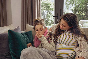 Eine schwangere Mutter sitzt mit ihrer Tochter im Grundschulalter auf einem Sofa. Die Tochter wirkt erk&auml;ltet. Sie isst eine Decke geh&uuml;llt, sieht fiebrig aus und hustet.