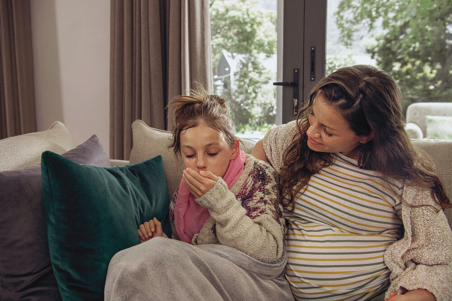 Eine schwangere Mutter sitzt mit ihrer Tochter im Grundschulalter auf einem Sofa. Die Tochter wirkt erkältet. Sie isst eine Decke gehüllt, sieht fiebrig aus und hustet.
