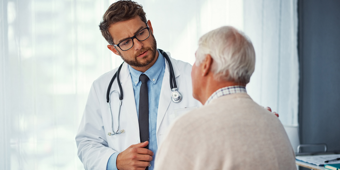 Foto: Ein junger Arzt im weißen Kittel spricht mit einem älteren Herrn, der von hinten zu sehen ist.