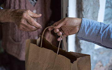 Close-Up: Eine Hand reicht von au&szlig;en eine Papiert&uuml;te einer anderen, &auml;lteren Hand ins Haus.