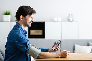 Foto_PM_Online-Coach Bluthochdruck mit neuen Funktionen Ein Mann sitzt mit einem Blutdruckmessgerät am Arm an einem Tisch und schaut auf sein Handy.