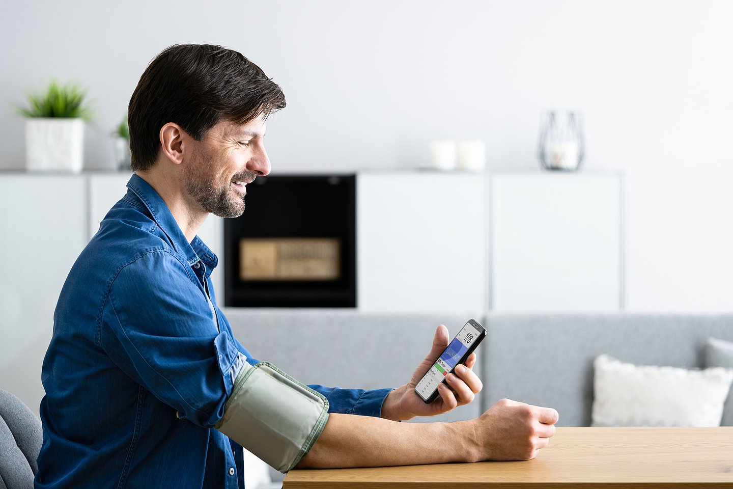 Ein Mann sitzt mit einem Blutdruckmessgerät am Arm an einem Tisch und schaut auf sein Handy.
