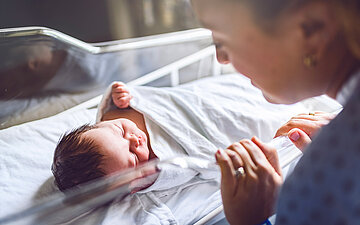 Eine Person beugt sich &uuml;ber ein schlafendes Neugeborenes und h&auml;lt seine Hand.