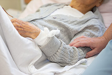 Ein &auml;lterer Mensch liegt in einem Bett. Am linken Handgelenk ist ein Verband sichtbar, der zum Teil von einer Strickjacke abgedeckt wird. Auf dem Oberarm liegt die Hand einer anderen Person, die nicht sichtbar ist.