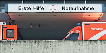 Notaufnahme Der Eingang zur Notaufnahme: Zwei Rettungsfahrzeuge stehen vor dem Einfang. Über dem Eingang steht auf dem Schild "Erste Hilfe" und "Notaufnahme".
