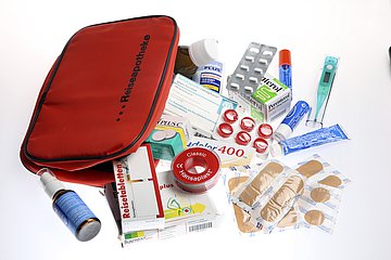 Reiseapotheke Bild zeigt ein rotes Täschechen mit der Aufschrift "Reiseapotheke". Drumherum verscheiedene Medikamente un dMediziprodukte wie Tabeltte, Nasenspray, Pflaster und Fieberthermometer.