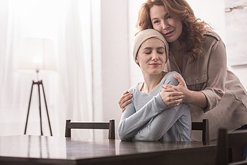 Ambulante Hospizdienste in Schleswig-Holstein Foto zeigt eine kranke junge Frau an einem Tisch sitzend, die von einer stehenden Frau hinter ihr an den Schultern gehalten wird.hinten