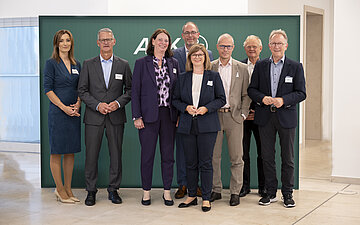 Das Foto zeigt ein Gruppenbild mit insgesamt acht Personen (drei Frauen, f&uuml;nf M&auml;nner). Auf der dunkelgr&uuml;nen Wand im Hintergrund ist schemenhaft das AOK-Logo zu sehen.