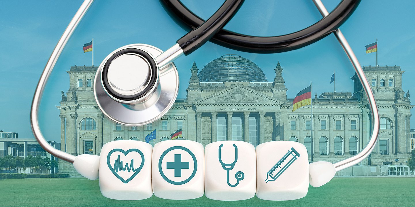 Foto des Bundestags im Sommer. Davor im Bild sind ein Stethoskop zu sehen und vier Würfel mit medizinischen Symbolen