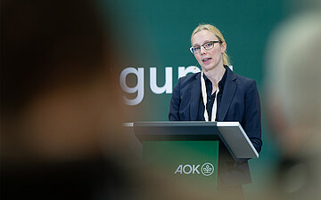 Anja Sch&ouml;dwell steht bei ihrem Vortrag auf der Selbsthilfe-Fachtagung des AOK-Bundesverbandes am 12.1.2024 in Berlin hinter einem Stehpult auf der B&uuml;hne.