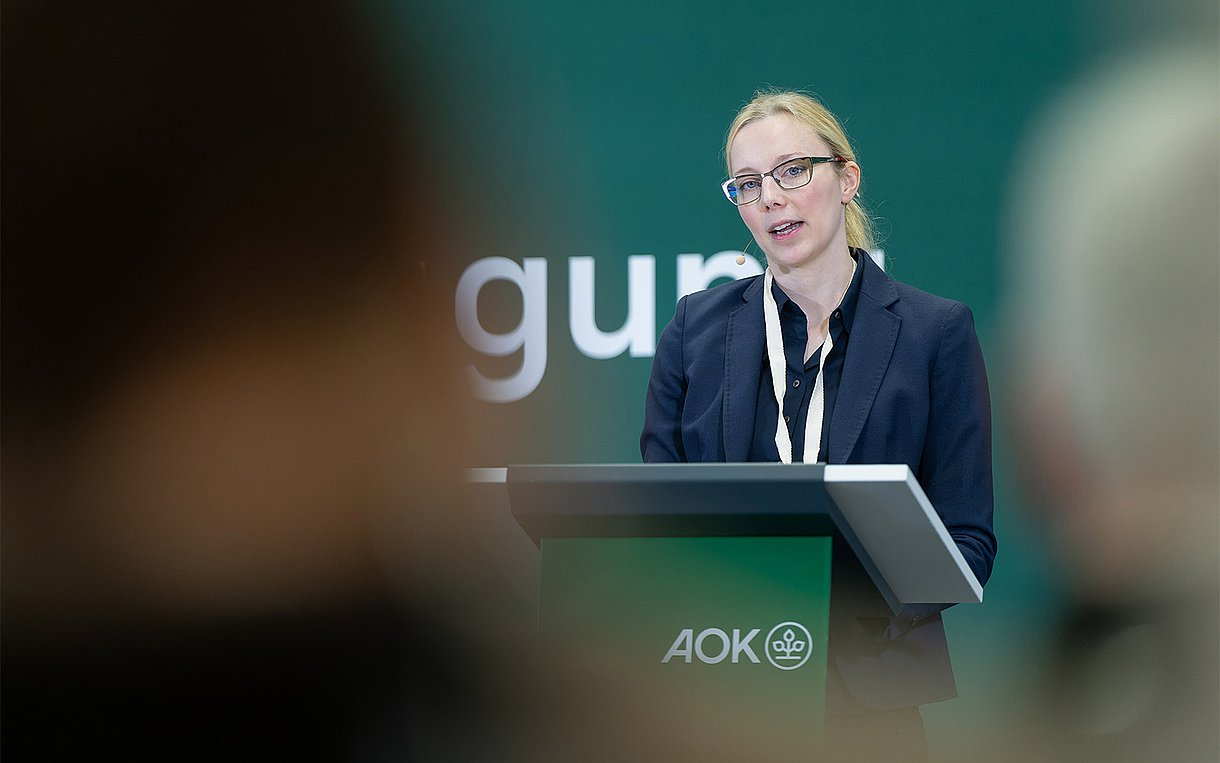 Anja Schödwell steht bei ihrem Vortrag auf der Selbsthilfe-Fachtagung des AOK-Bundesverbandes am 12.1.2024 in Berlin hinter einem Stehpult auf der Bühne.