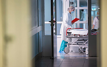 Foto: Zwei Personen in medizinischer Schutzkleidung im Flur eines Krankenhauses an einem Krankenbett.